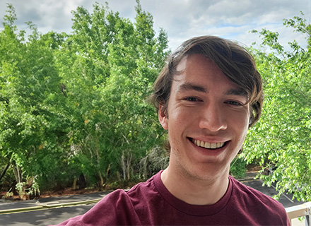 matt, wearing a maroon tee and standing in front of a group of trees, smiles